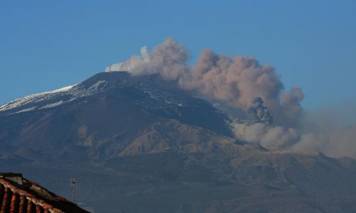 Vista del vulcano Etna con una colata lavica che solleva fumo e cenere, con tetti in primo piano