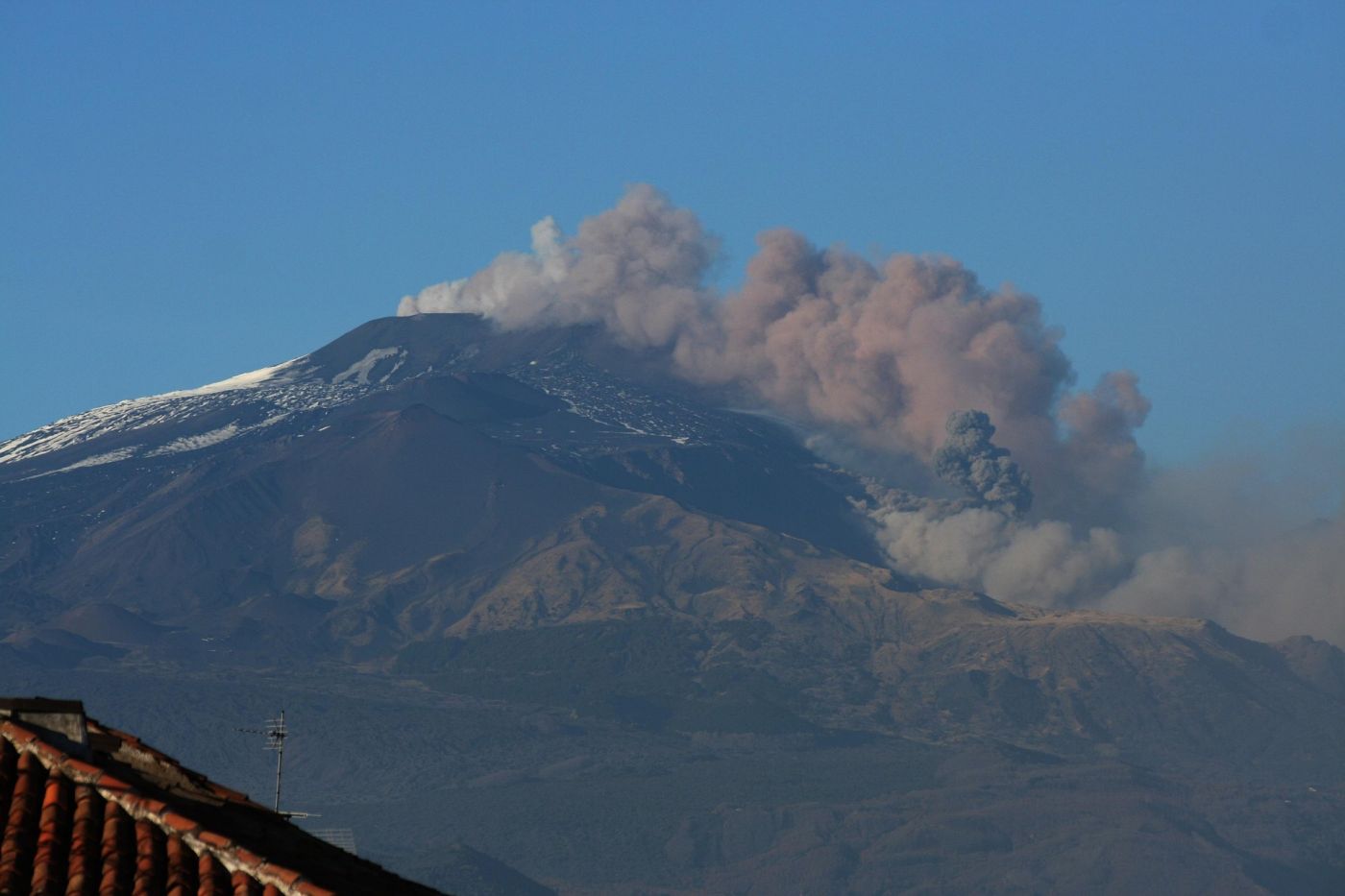 Vista del vulcano Etna con una colata lavica che solleva fumo e cenere, con tetti in primo piano