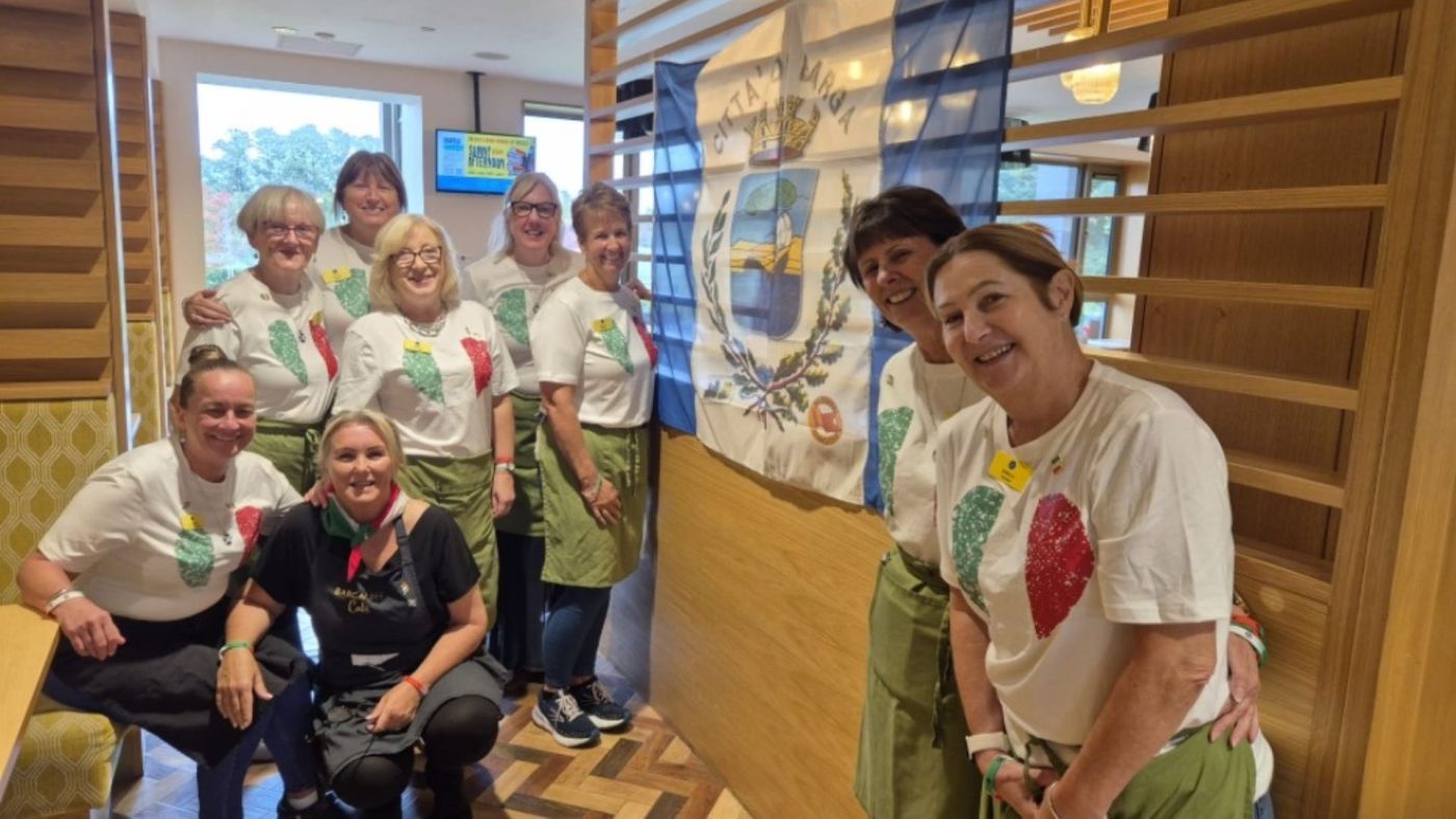 Gruppo di volontarie con magliette tricolori e grembiuli verdi durante la Festa Italiana, sorridenti di fronte a una bandiera comunale