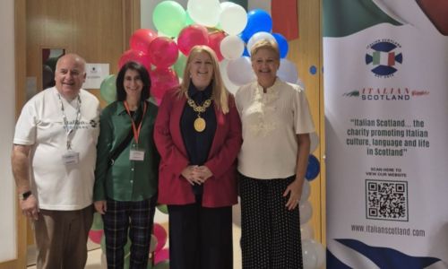 Quattro persone posano sorridenti di fronte a palloncini colorati e al banner dell’associazione Italian Scotland durante la Festa Italiana.