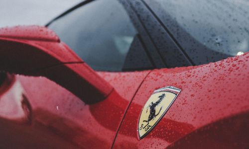 Particolare di una Ferrari rossa sotto la pioggia con il logo del Cavallino Rampante in primo piano